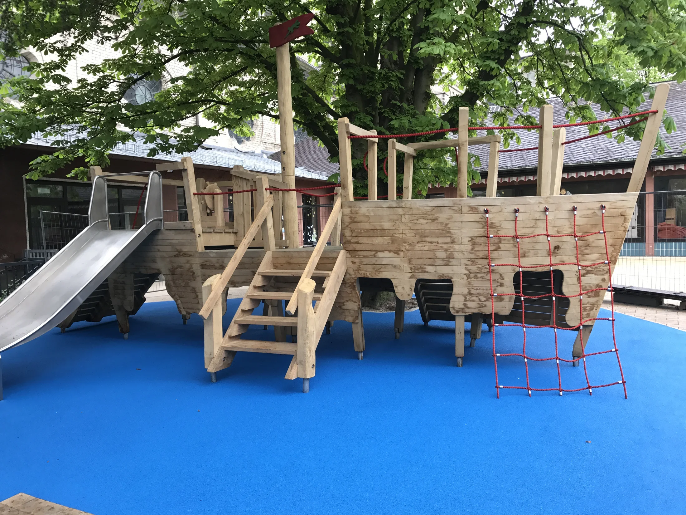 Blaue fugenlose Fallschutzfläche unter einem Spielschiff aus Holz mit Rutsche und Kletternetz